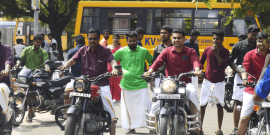 A group of men dressed in traditional attire and colored shirts are sitting on motorcycles, ready to start a ride. They are outdoors with others standing around. A school bus is parked in the background, and a sign for KVIM Business School is visible.