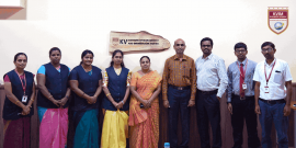 A group of nine people stands in a row, posing for a photo in an indoor setting. They are dressed in business casual and traditional attire. A wooden plaque on the wall behind reads "KV Institute Of Management And Information Studies." A logo is visible in the top right corner.