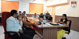 A group of people is sitting around a conference table engaging in a discussion. Some participants are looking at a laptop, while others are speaking. The room has orange window blinds, a large pedestal fan, water bottles on the table, and a KVIM Business School logo on the wall.