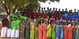 A large group of people, consisting of young men and women along with a few older individuals, stand posed for a photo outdoors. The women wear colorful sarees, and some men wear traditional attire. They stand under a tree with a sign "KVIM Business School.