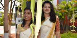 Two women wearing traditional white and gold sarees stand outdoors, smiling. They are partially obscured by hanging decorations made of palm leaves. The background includes greenery, a tree, and part of a building.