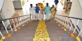 A group of people, including three men and one woman in traditional attire, carry a decorative object up stairs adorned with flower petals. Another woman, also in traditional attire, waits at the top of the stairs. The setting appears festive.