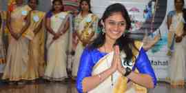 A young woman in a blue and white traditional outfit smiles and holds her hands in a namaste gesture. She is standing in front of a group of women similarly dressed in traditional attire. The background is decorated with colorful balloons and a backdrop showing an event name.