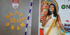 A woman dressed in traditional attire is speaking into a microphone at an event. The backdrop has colorful decorations, the phrase "Happy Onam 2K19," and the KVIM Business School logo. A decorative Onam-themed mural is visible in the background.