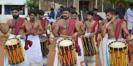 A group of men dressed in traditional attire, bare-chested with white dhotis and colorful sashes, are playing drums outdoors. They are engaged in a lively performance, with some men wearing jewelry and displaying intense expressions. Trees and a building are in the background.