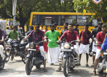 A group of men dressed in traditional attire and colored shirts are sitting on motorcycles, ready to start a ride. They are outdoors with others standing around. A school bus is parked in the background, and a sign for KVIM Business School is visible.