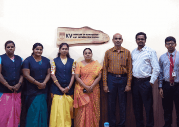 A group of nine people stands in a row, posing for a photo in an indoor setting. They are dressed in business casual and traditional attire. A wooden plaque on the wall behind reads "KV Institute Of Management And Information Studies." A logo is visible in the top right corner.