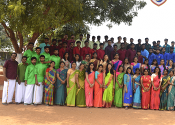 A large group of people, consisting of young men and women along with a few older individuals, stand posed for a photo outdoors. The women wear colorful sarees, and some men wear traditional attire. They stand under a tree with a sign "KVIM Business School.