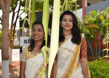 Two women wearing traditional white and gold sarees stand outdoors, smiling. They are partially obscured by hanging decorations made of palm leaves. The background includes greenery, a tree, and part of a building.