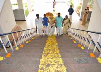 A group of people, including three men and one woman in traditional attire, carry a decorative object up stairs adorned with flower petals. Another woman, also in traditional attire, waits at the top of the stairs. The setting appears festive.