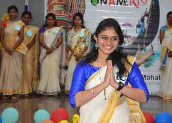 A young woman in a blue and white traditional outfit smiles and holds her hands in a namaste gesture. She is standing in front of a group of women similarly dressed in traditional attire. The background is decorated with colorful balloons and a backdrop showing an event name.