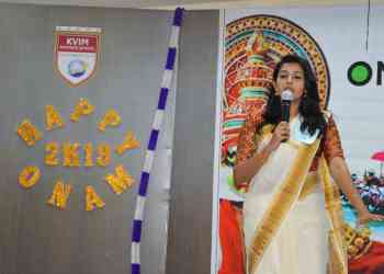 A woman dressed in traditional attire is speaking into a microphone at an event. The backdrop has colorful decorations, the phrase "Happy Onam 2K19," and the KVIM Business School logo. A decorative Onam-themed mural is visible in the background.