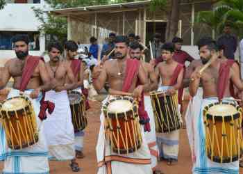 A group of men dressed in traditional attire, bare-chested with white dhotis and colorful sashes, are playing drums outdoors. They are engaged in a lively performance, with some men wearing jewelry and displaying intense expressions. Trees and a building are in the background.