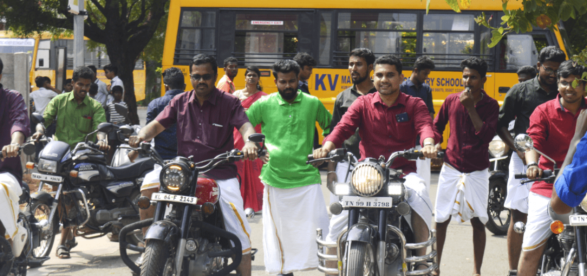 A group of men dressed in traditional attire and colored shirts are sitting on motorcycles, ready to start a ride. They are outdoors with others standing around. A school bus is parked in the background, and a sign for KVIM Business School is visible.