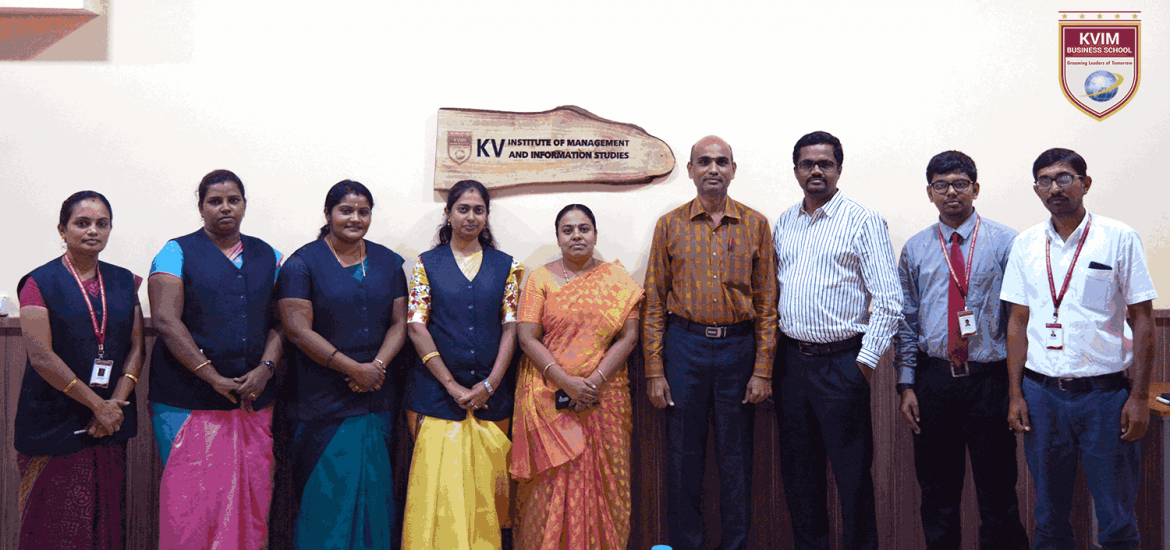 A group of nine people stands in a row, posing for a photo in an indoor setting. They are dressed in business casual and traditional attire. A wooden plaque on the wall behind reads "KV Institute Of Management And Information Studies." A logo is visible in the top right corner.