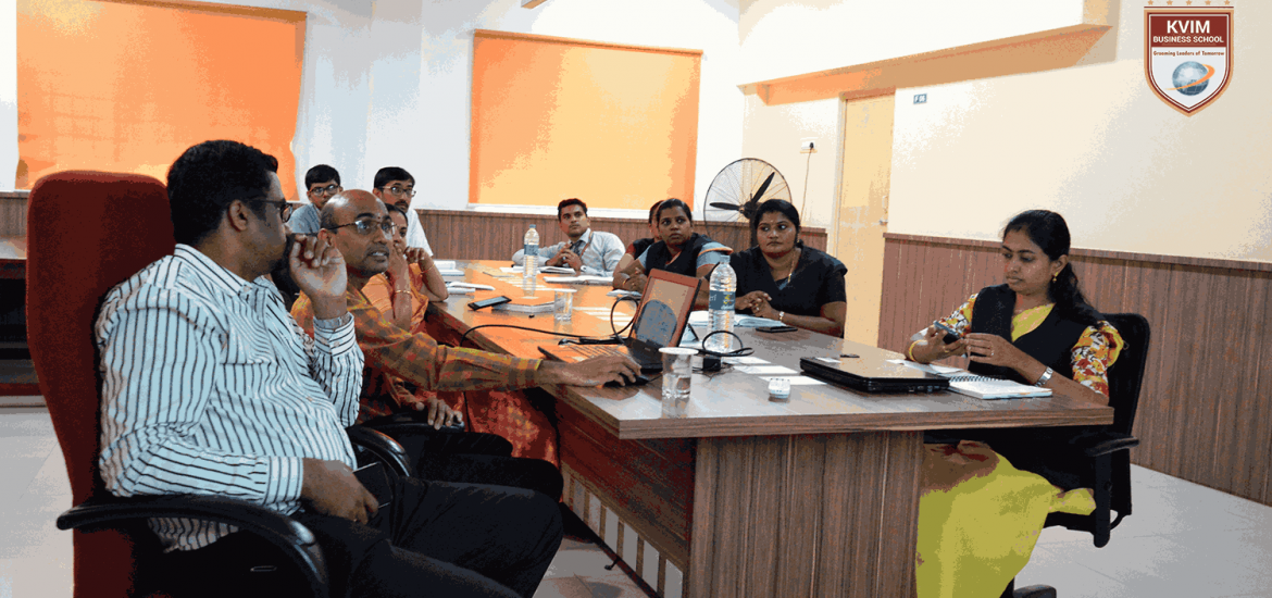 A group of people is sitting around a conference table engaging in a discussion. Some participants are looking at a laptop, while others are speaking. The room has orange window blinds, a large pedestal fan, water bottles on the table, and a KVIM Business School logo on the wall.