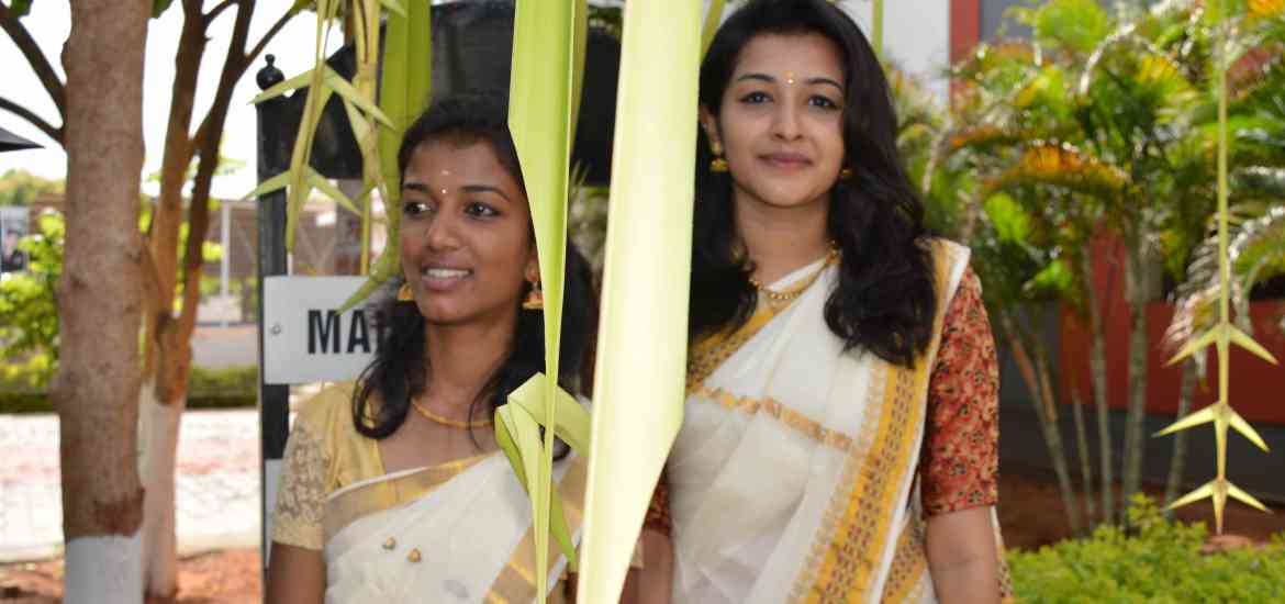 Two women wearing traditional white and gold sarees stand outdoors, smiling. They are partially obscured by hanging decorations made of palm leaves. The background includes greenery, a tree, and part of a building.