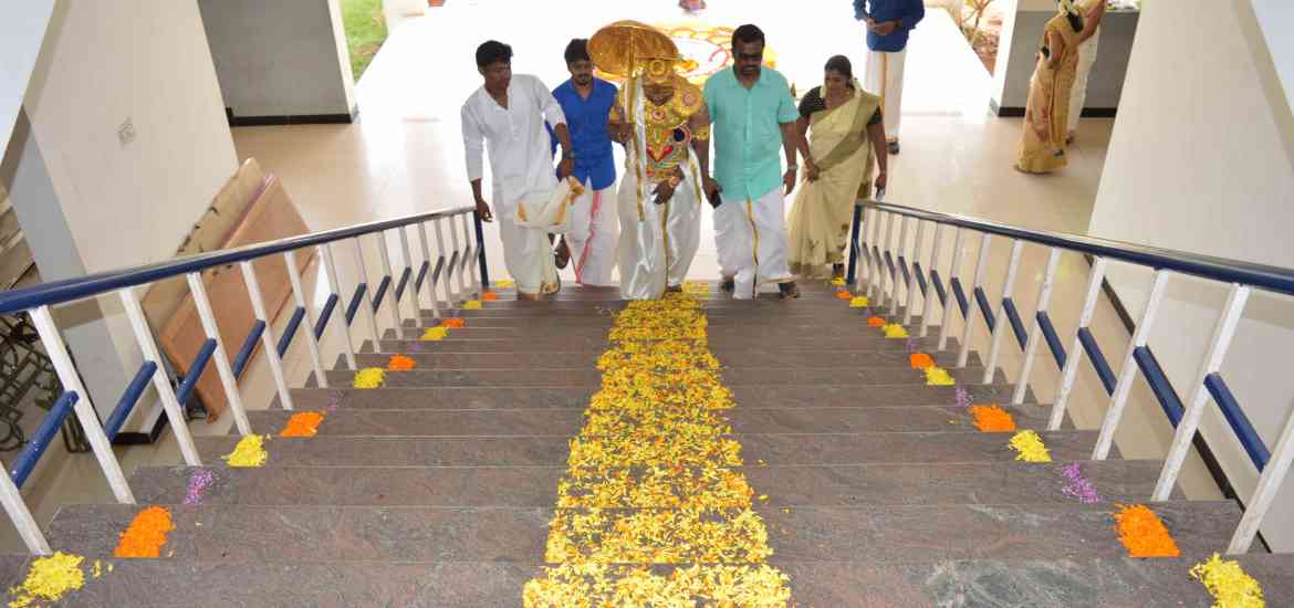 A group of people, including three men and one woman in traditional attire, carry a decorative object up stairs adorned with flower petals. Another woman, also in traditional attire, waits at the top of the stairs. The setting appears festive.