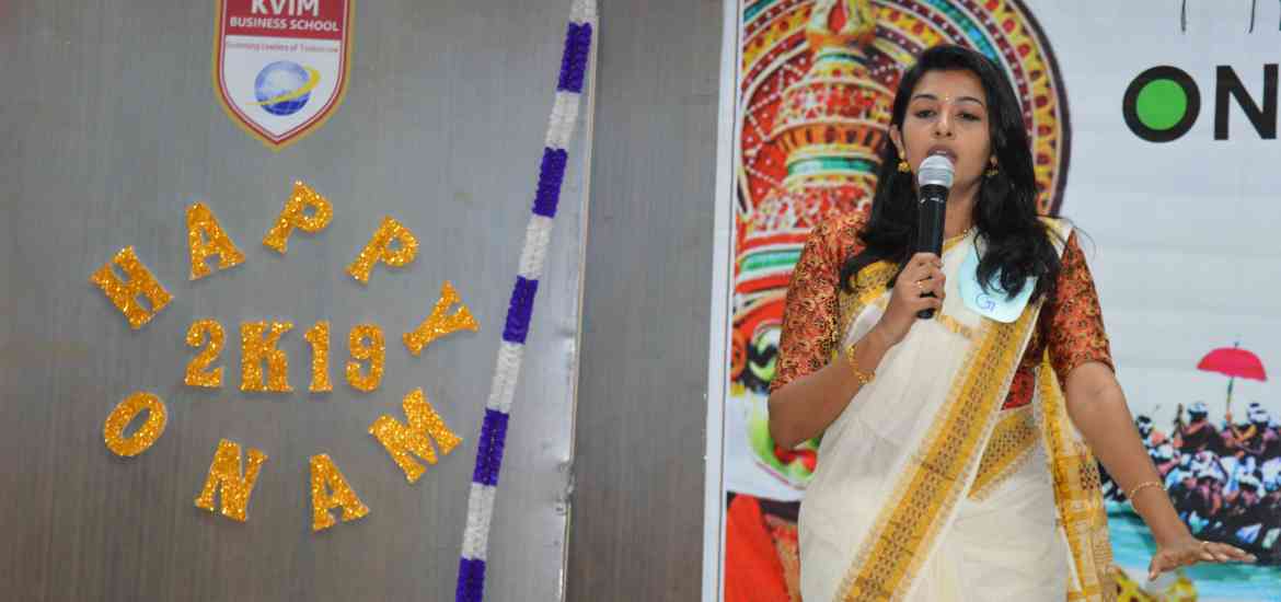 A woman dressed in traditional attire is speaking into a microphone at an event. The backdrop has colorful decorations, the phrase "Happy Onam 2K19," and the KVIM Business School logo. A decorative Onam-themed mural is visible in the background.