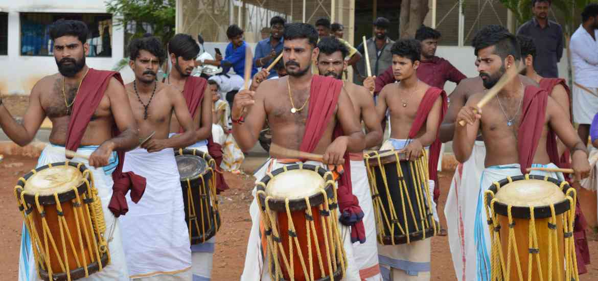 A group of men dressed in traditional attire, bare-chested with white dhotis and colorful sashes, are playing drums outdoors. They are engaged in a lively performance, with some men wearing jewelry and displaying intense expressions. Trees and a building are in the background.