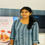 A young woman with dark hair tied back stands indoors, smiling. She is wearing a blue striped kurti. Behind her, there is a banner partially visible with images of food and the text "local treat". A black television screen is also seen in the background.